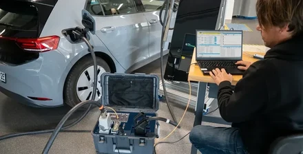 Geöffneter EVCA Interop-Koffer zwischen einem realen Elektrofahrzeug und einer Ladestation, während ein Ingenieur die synchronisierte Sitzung analysiert